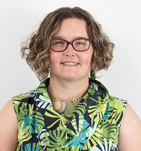 Headshot of Bex Dunn. Bex wears a palm-leaf pattern shirt with green black and teal colors. She has dark blond hair and oval shaped glasses.