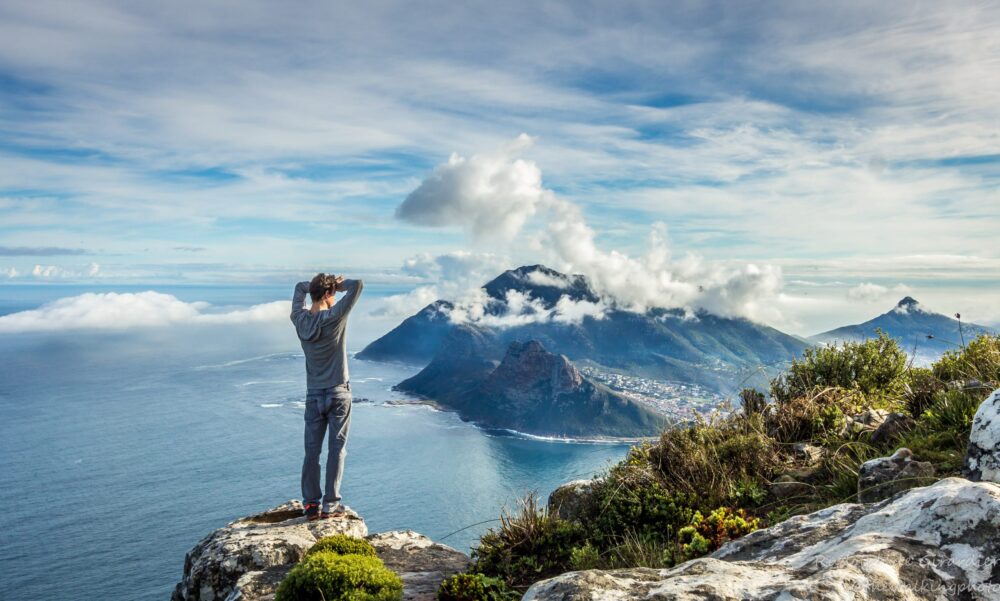 Man standing on rock cliff with his hands on his head looking at the ocean and mountain tops of Table Mountain National Park. Source: South Africa National Parks