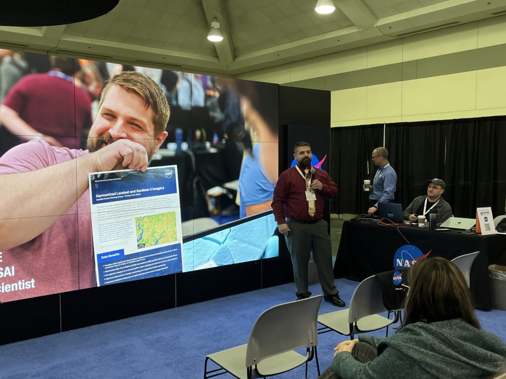 Mike Taylor in front of the NASA hyperwall. He is wearing dark pants and a maroon-colored shirt. A large photo of him holding up an information sheet about Harmonized Landsat Sentinel-2 data is shown on the large screen.