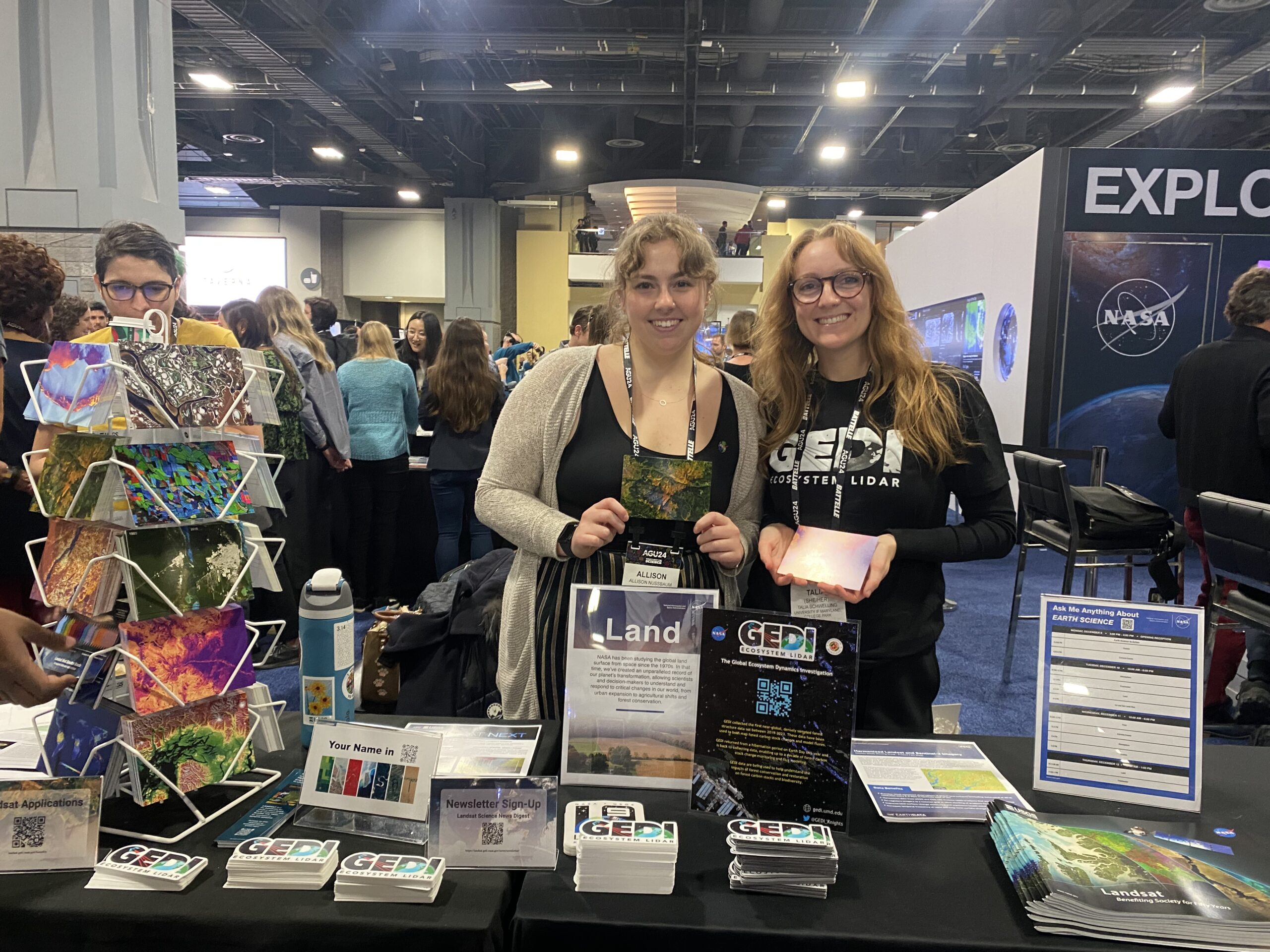 Landsat outreach specialist Allison Nussbaum and GEDI/UMD Faculty Specialist Talia Schwelling hold Landsat/GEDI postcards at the NASA exhibit booth at AGU24.