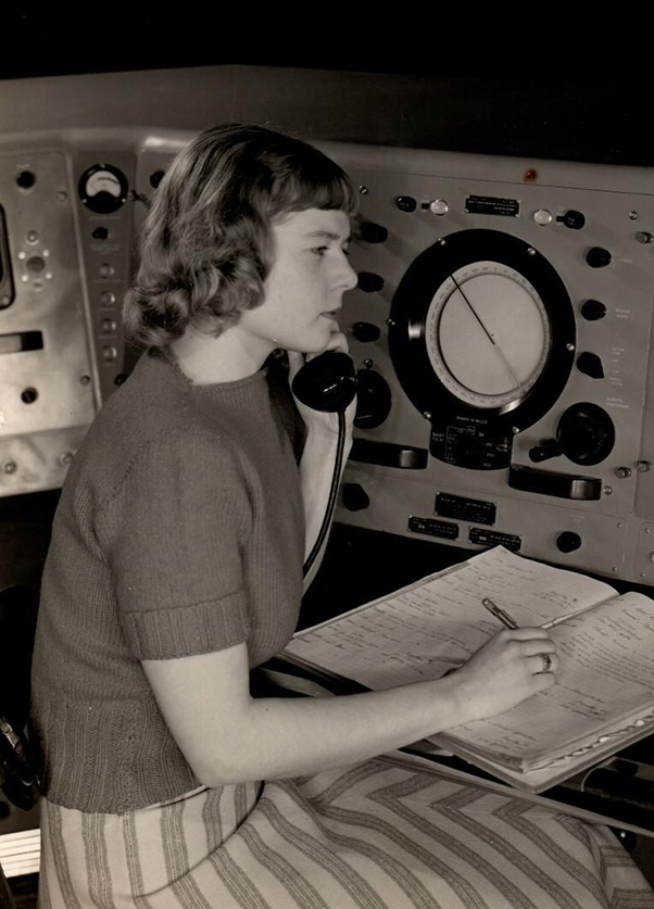 A black and white photo of Virginia Norwood on a telephone in front of a radar system writing in a notebook.