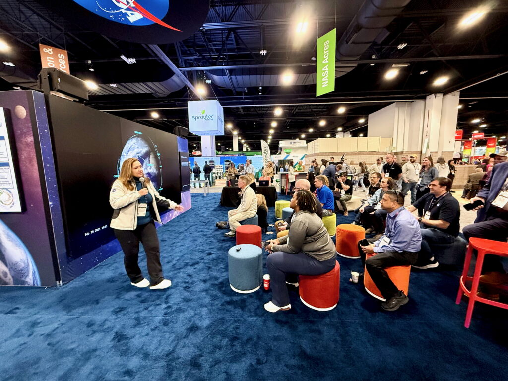 NASA Earth Science Division Director Karen St. Germain speaks in front of a large screen to a group of listeners on stools.