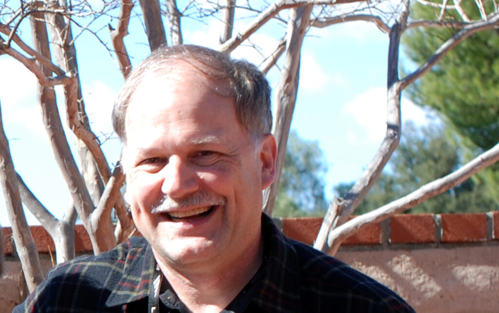 A headshot of Brian Markham. Tree branches can be seen in the background; Brian is smiling.