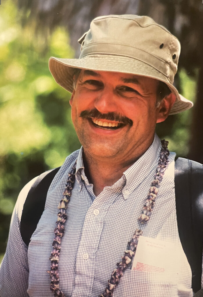 Brian wearing a sunhat, backpack, and beaded necklace in Hawaii.