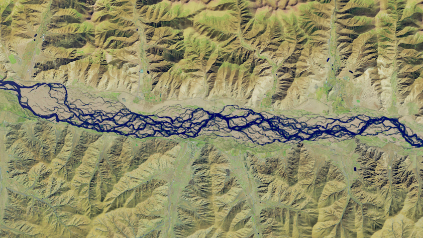 A false-color satellite image shows numerous intertwining channels of blue water spanning the middle of the scene from left to right. The river is bordered on the top and bottom by mountainous terrain, which appears tan and light green.